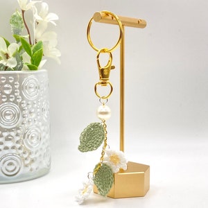 May include: A white crocheted flower keychain with green leaves and a pearl charm. The keychain is attached to a gold metal key ring and is displayed on a gold stand.