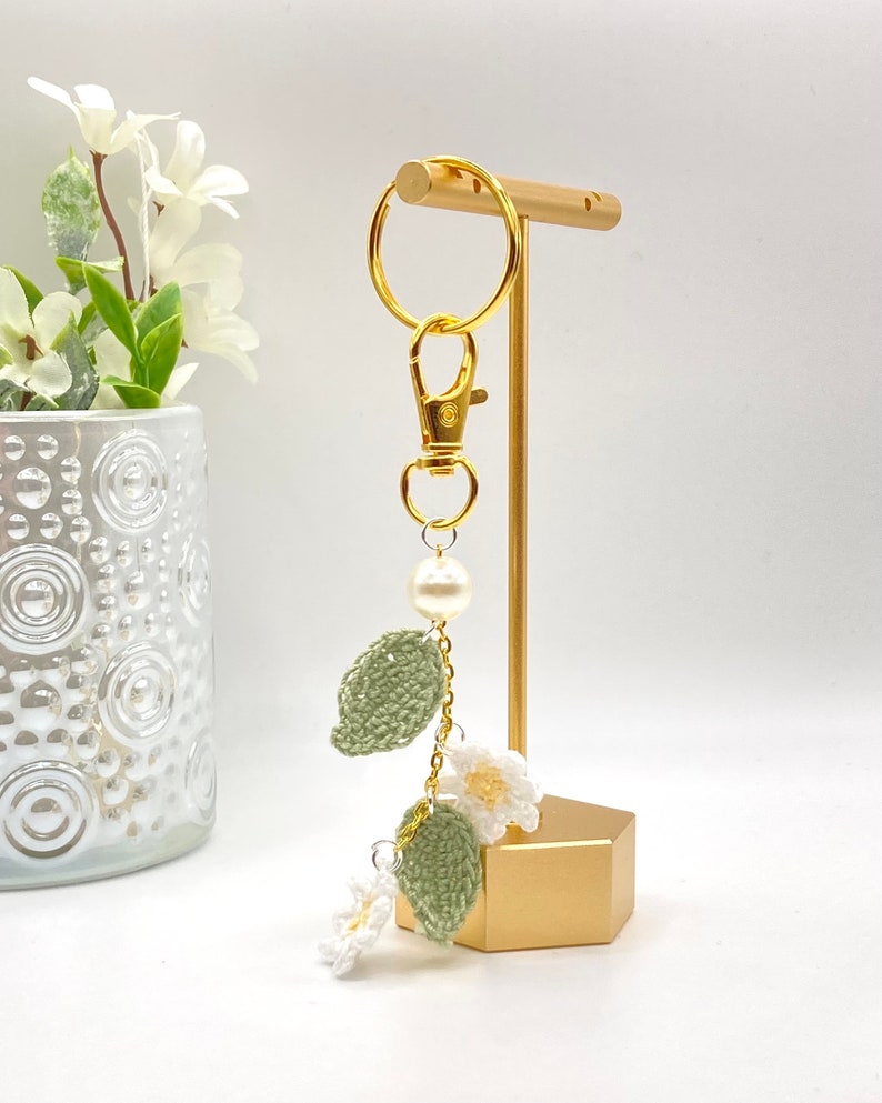 May include: A white crocheted flower keychain with green leaves and a pearl charm. The keychain is attached to a gold metal key ring and is displayed on a gold stand.