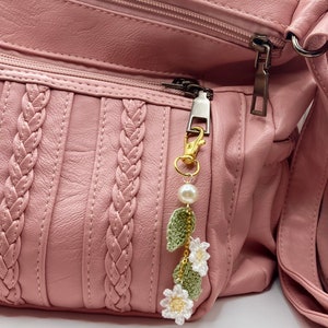 May include: A pink leather purse with a braided design and a gold zipper pull. A white crocheted flower charm with green leaves and a pearl hangs from the zipper pull.
