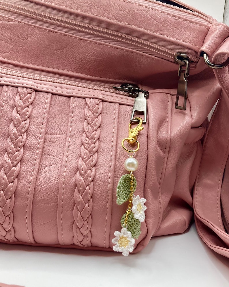 May include: A pink leather purse with a braided design and a gold zipper pull. A white crocheted flower charm with green leaves and a pearl hangs from the zipper pull.