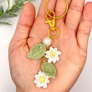 May include: A crocheted keychain with white flowers and green leaves, accented with a pearl and a gold key ring.