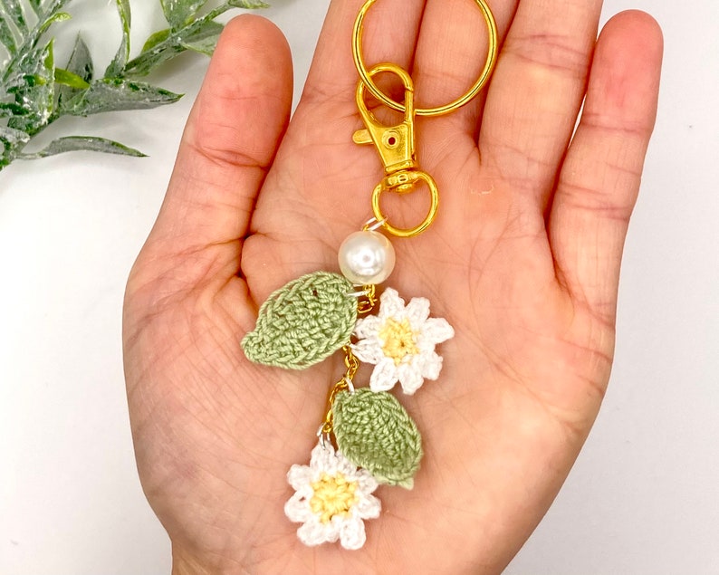 May include: A crocheted keychain with white flowers and green leaves, accented with a pearl and a gold key ring.