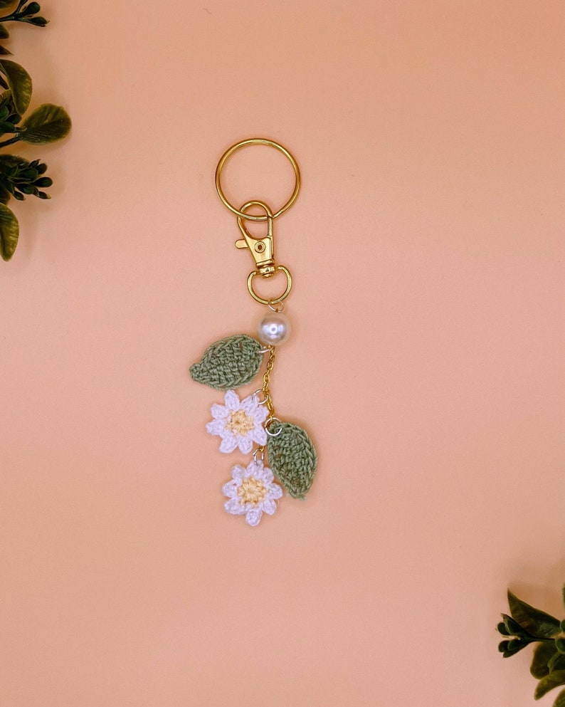 May include: A white crocheted flower keychain with green leaves and a gold key ring. The keychain has a pearl bead accent.