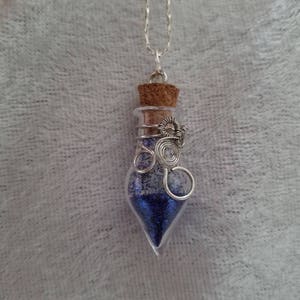 Puede incluir: Un collar de cadena plateada con un pequeño colgante de botella de vidrio lleno de purpurina azul. La botella tiene un tapón de corcho y está envuelta con alambre plateado.