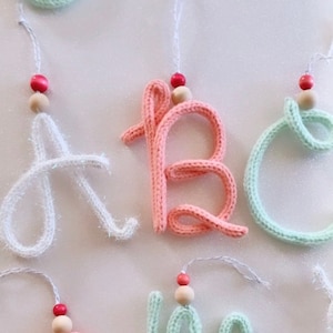 May include: Knitted yarn letters with wooden beads and red beads, forming the word "ABC" and "LM", in white, pink, and green.