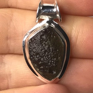 May include: A teardrop-shaped pendant featuring a dark brown, textured moldavite stone set in a polished silver frame. The pendant includes a silver bail at the top for attachment to a chain, showcasing a contrast between the rough stone and metal.