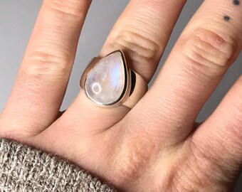 Könnte beinhalten: Ein silberner Ring mit einem tropfenförmigen Mondstein. Der Ring befindet sich an einem Finger.