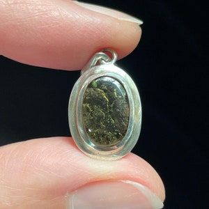 Puede incluir: Un colgante ovalado de plata con una piedra verde oscuro y dorada engastada en su interior.