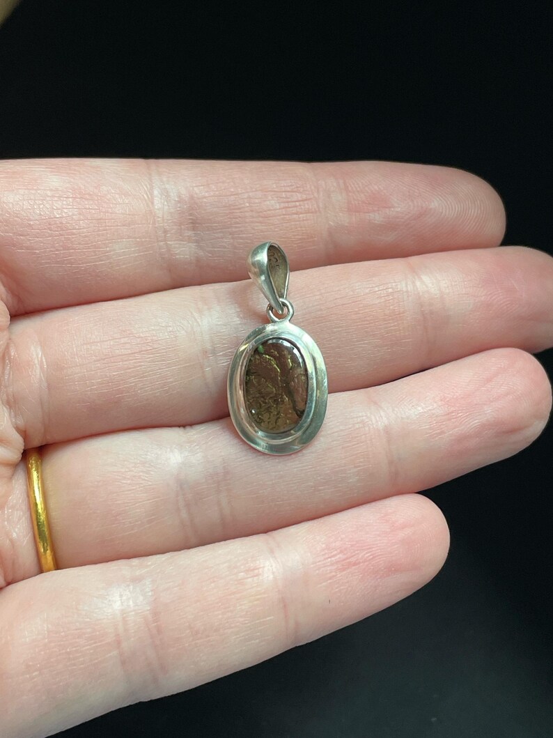 Puede incluir: Un colgante de plata con una piedra preciosa ovalada de color marr&oacute;n y verde engastada en un engaste de plata.