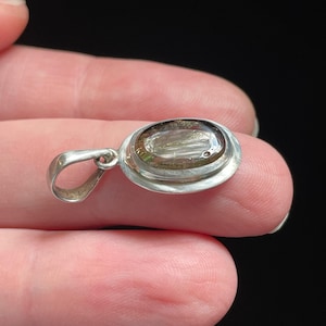 Puede incluir: Un colgante de plata con una piedra ovalada transparente con motas marrones y plateadas. La piedra est&aacute; engastada en un bisel de plata con un asa de plata.