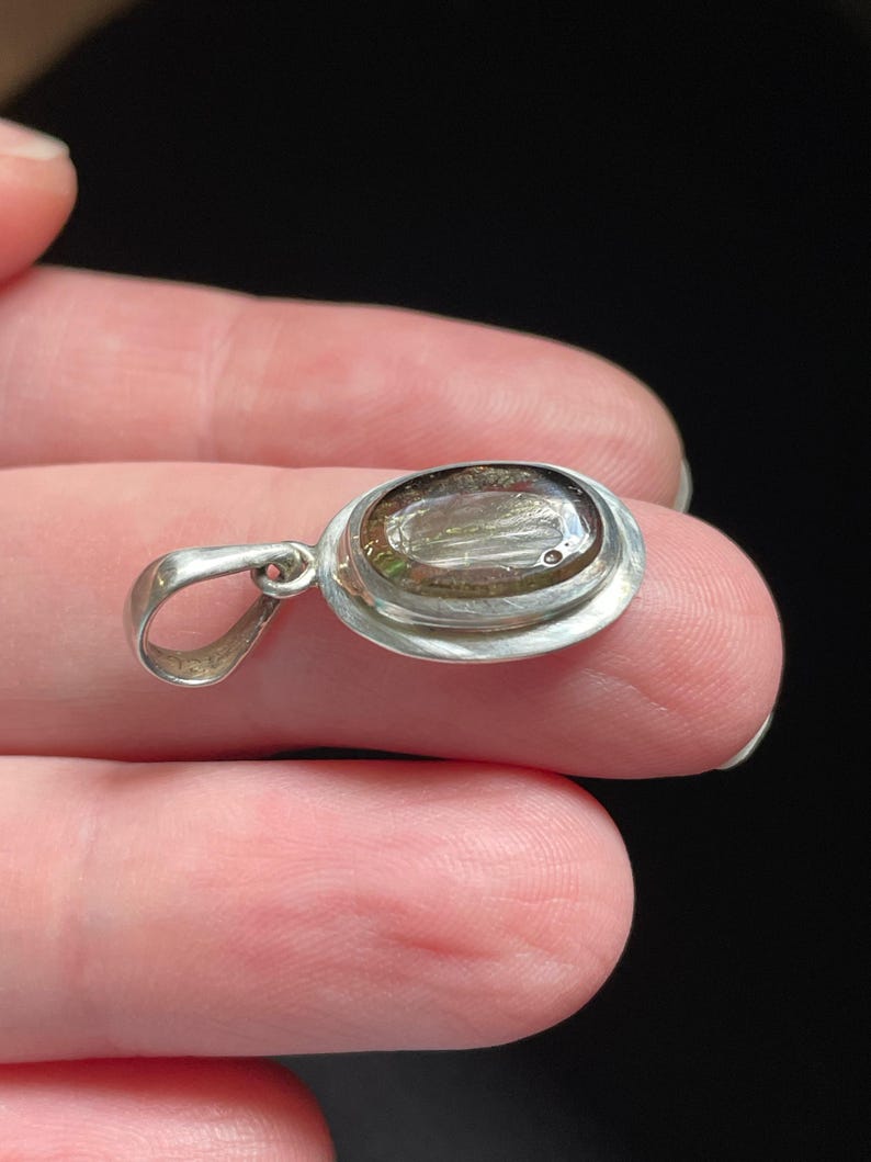Puede incluir: Un colgante de plata con una piedra ovalada transparente con motas marrones y plateadas. La piedra est&aacute; engastada en un bisel de plata con un asa de plata.