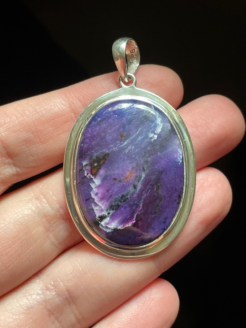 May include: A silver pendant with a purple and white stone set in an oval bezel. The stone has a swirling pattern and is surrounded by a silver border. The pendant has a silver bail with the number "925" stamped on it.