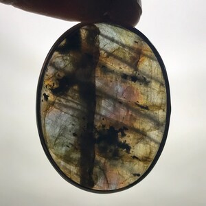 May include: Oval-shaped labradorite gemstone pendant with a silver bezel setting. The stone has a dark gray, black, and brown color with iridescent flashes of blue, green, and gold.