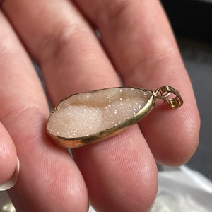 Puede incluir: Un colgante dorado con una piedra ovalada blanca y brillante.