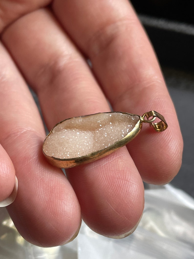 Puede incluir: Un colgante dorado con una piedra ovalada blanca y brillante.