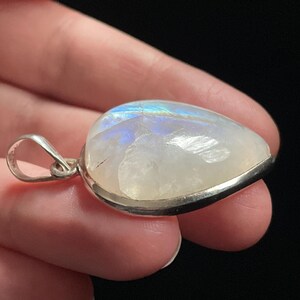May include: A teardrop-shaped moonstone pendant with a silver setting. The stone has a milky white color with iridescent blue and green flashes.