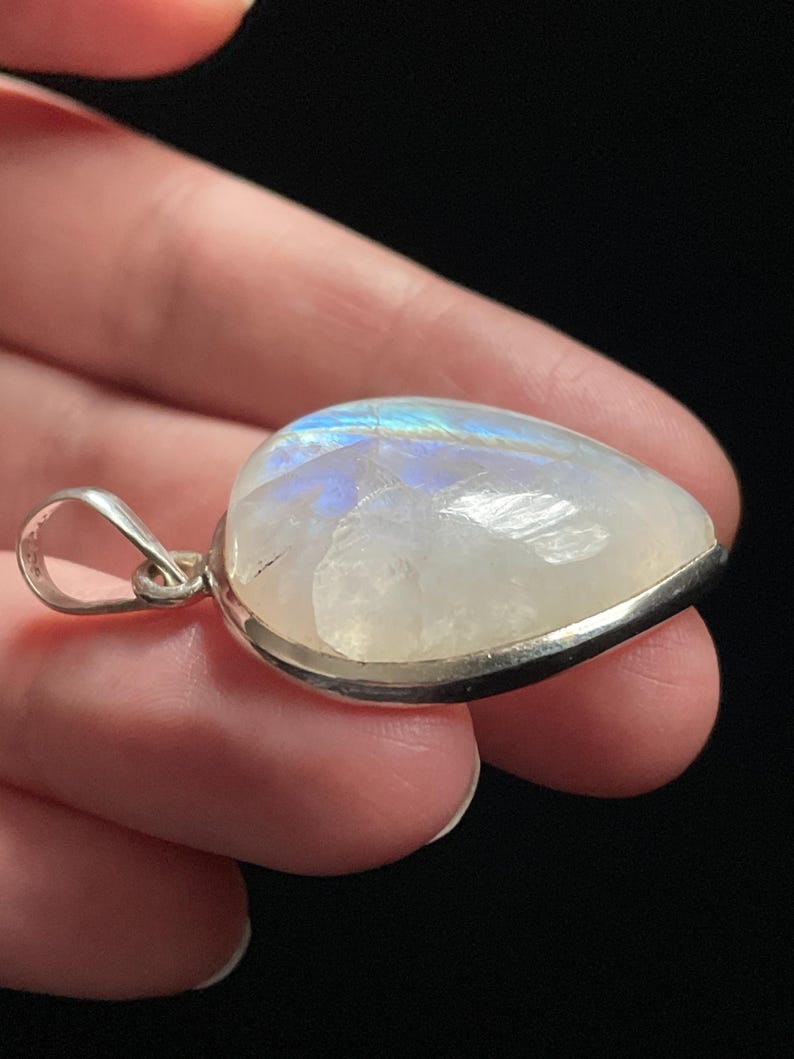 May include: A teardrop-shaped moonstone pendant with a silver setting. The stone has a milky white color with iridescent blue and green flashes.