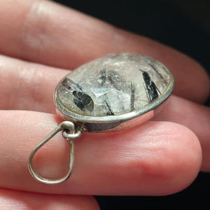 May include: A silver pendant with a clear quartz stone containing black tourmaline inclusions. The pendant has a simple loop bail.