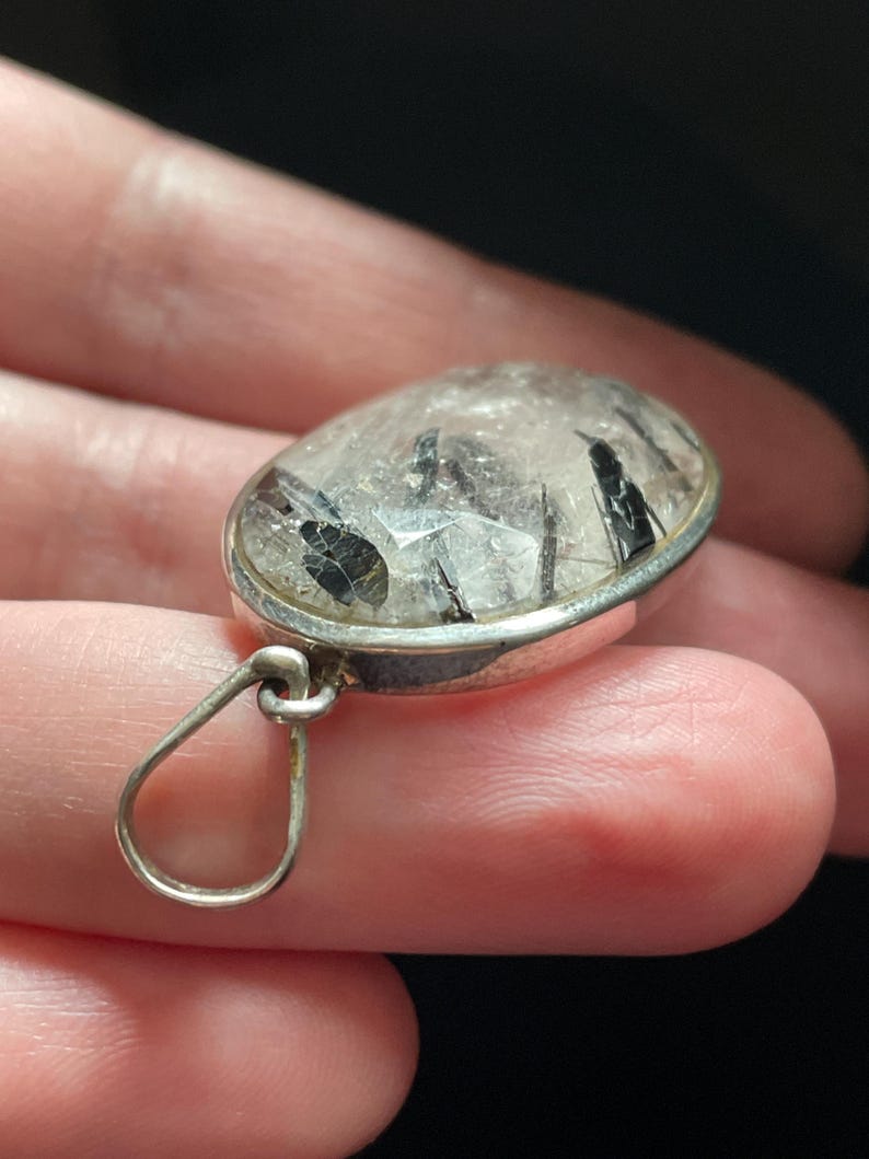 May include: A silver pendant with a clear quartz stone containing black tourmaline inclusions. The pendant has a simple loop bail.