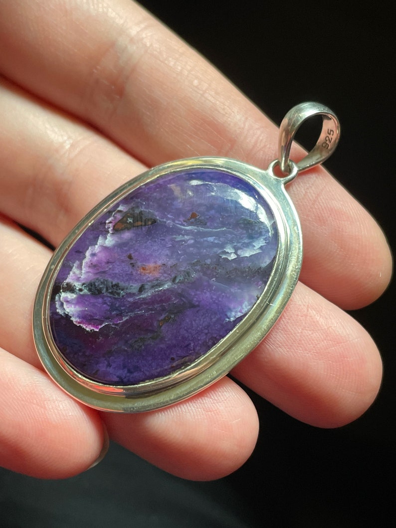 May include: A silver pendant with a purple and white stone set in a silver oval frame. The pendant has a silver bail with the number "925" stamped on it.