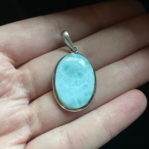 Puede incluir: Un colgante de plata con una piedra ovalada de color azul claro con un patr&oacute;n de remolino. El colgante est&aacute; en una mano.