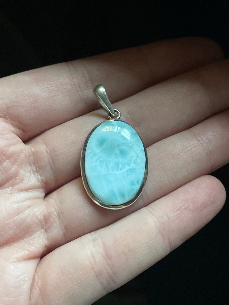 Puede incluir: Un colgante de plata con una piedra ovalada de color azul claro con un patr&oacute;n de remolino. El colgante est&aacute; en una mano.