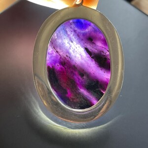 May include: A silver oval pendant with a purple and white agate stone. The stone has a swirling pattern and is set in a silver frame.