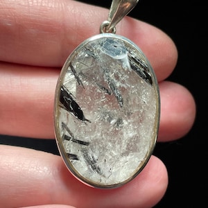 May include: Silver pendant featuring a clear quartz crystal with black tourmaline inclusions. The pendant is oval shaped and has a smooth, polished finish.