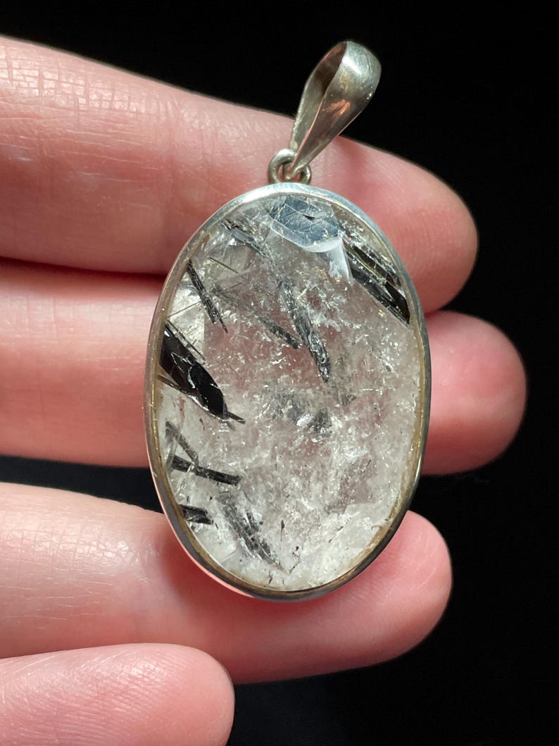 May include: Silver pendant featuring a clear quartz crystal with black tourmaline inclusions. The pendant is oval shaped and has a smooth, polished finish.