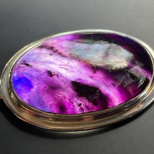 May include: A silver oval-shaped pendant with a purple and blue agate stone set in the center. The stone has a swirling pattern of colors.