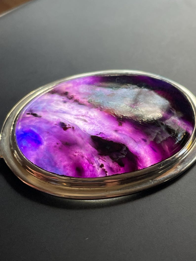 May include: A silver oval-shaped pendant with a purple and blue agate stone set in the center. The stone has a swirling pattern of colors.