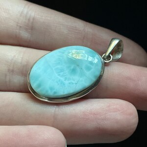 Puede incluir: Un colgante de plata con una piedra ovalada de color azul claro. La piedra tiene un patr&oacute;n de m&aacute;rmol y un borde blanco.