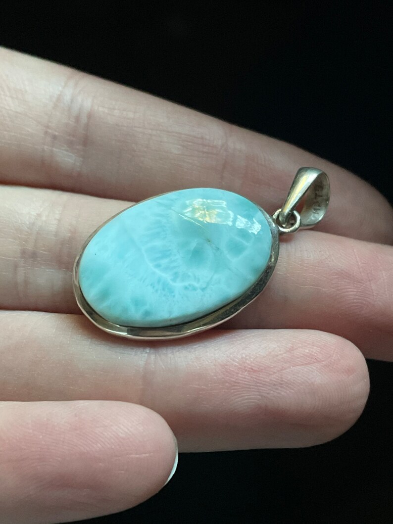 Puede incluir: Un colgante de plata con una piedra ovalada de color azul claro. La piedra tiene un patr&oacute;n de m&aacute;rmol y un borde blanco.