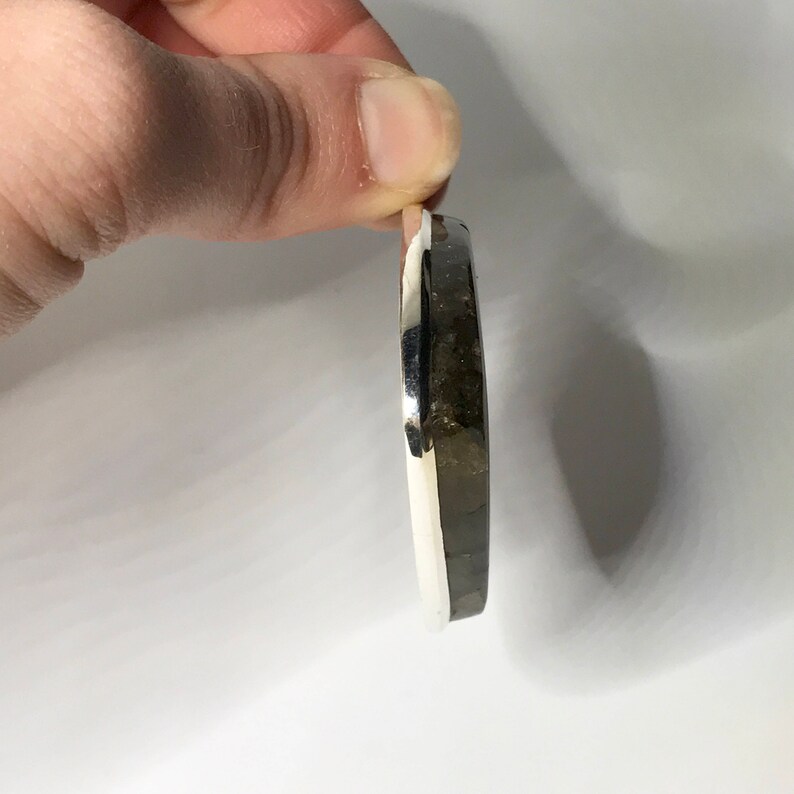 K&ouml;nnte beinhalten: Ein silberner Ring mit einem dunkelgrauen Stein, der in der Mitte eingesetzt ist. Der Ring wird von der Seite gezeigt, wobei der Stein zum Betrachter zeigt.