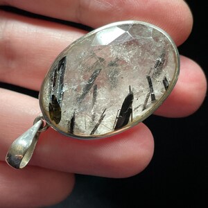 May include: A silver pendant featuring a clear quartz crystal with black tourmaline inclusions. The crystal is oval-shaped and has a faceted cut.