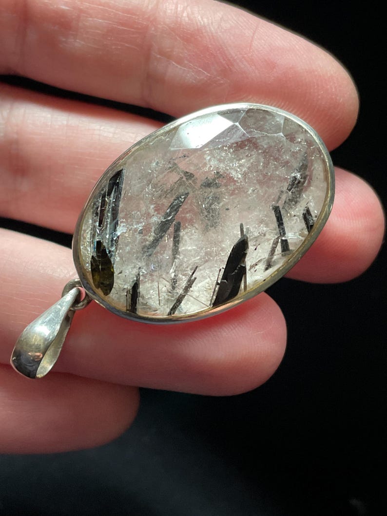 May include: A silver pendant featuring a clear quartz crystal with black tourmaline inclusions. The crystal is oval-shaped and has a faceted cut.