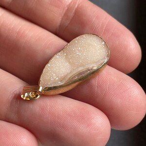 Puede incluir: Un colgante en forma de l&aacute;grima blanco y dorado con una textura brillante, similar a un cristal. El colgante tiene un aro de oro para sujetarlo a una cadena.