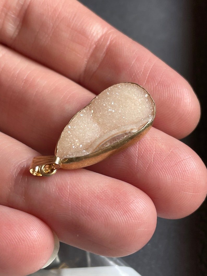 Puede incluir: Un colgante en forma de l&aacute;grima blanco y dorado con una textura brillante, similar a un cristal. El colgante tiene un aro de oro para sujetarlo a una cadena.