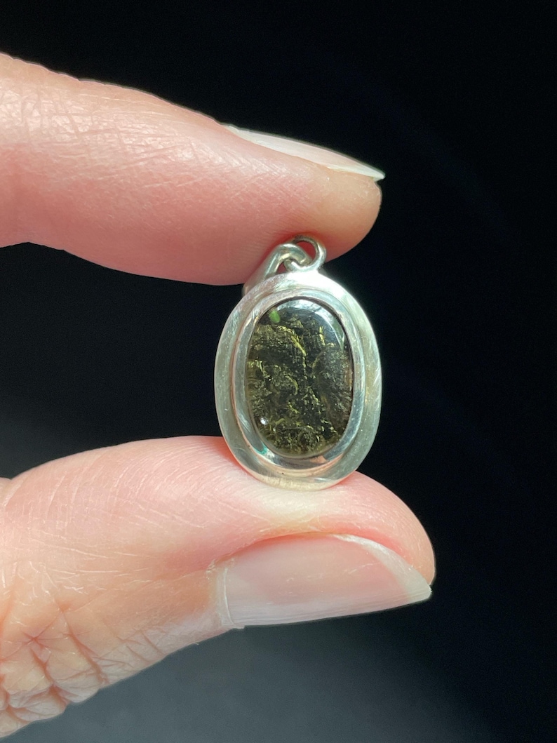 Puede incluir: Un colgante ovalado de plata con una piedra verde oscuro y dorada engastada en su interior.
