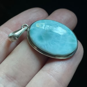 Puede incluir: Un colgante de plata con una piedra ovalada de color azul claro. La piedra tiene un patr&oacute;n de remolino y un borde blanco.