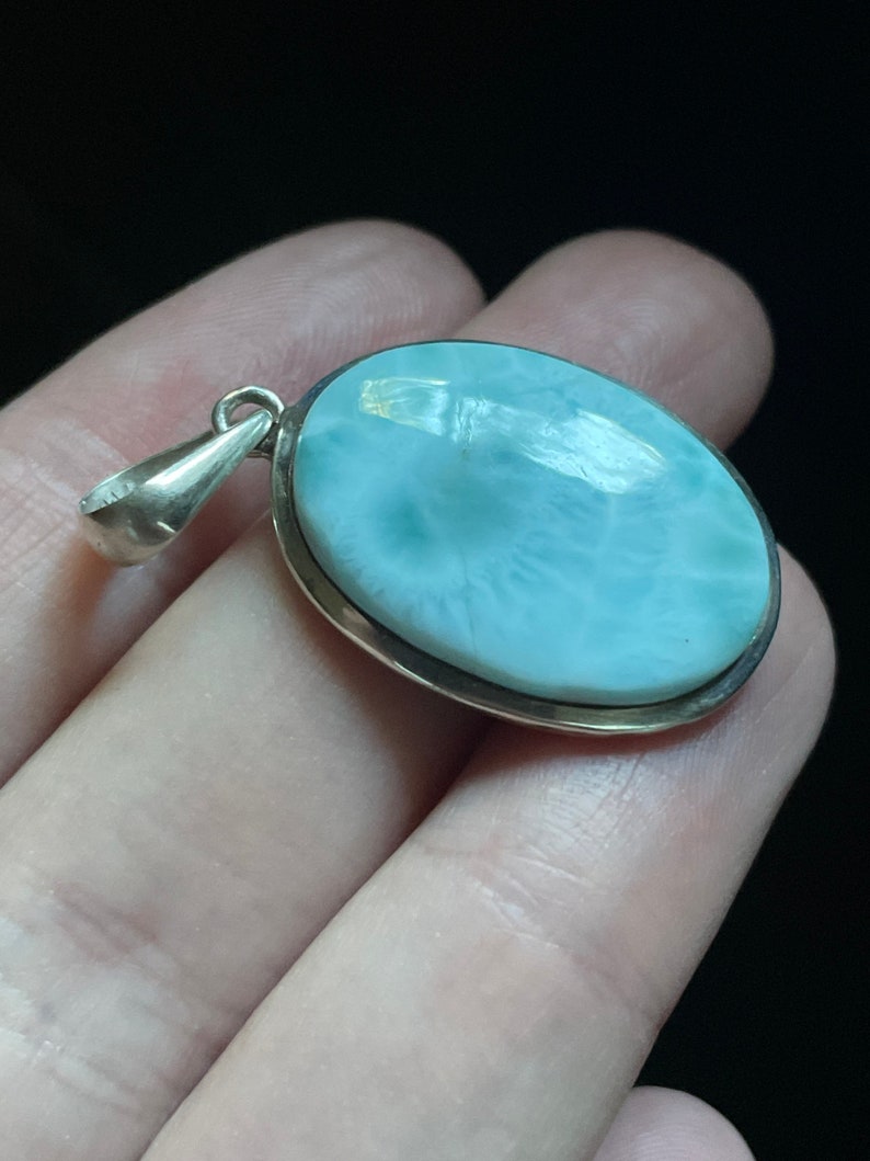 Puede incluir: Un colgante de plata con una piedra ovalada de color azul claro. La piedra tiene un patr&oacute;n de remolino y un borde blanco.