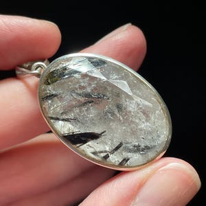 May include: A silver pendant featuring a clear quartz crystal with black tourmaline inclusions. The pendant is oval shaped and has a faceted design.