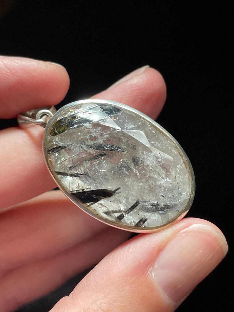 May include: A silver pendant featuring a clear quartz crystal with black tourmaline inclusions. The pendant is oval shaped and has a faceted design.