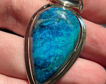 Può includere: Un ciondolo a forma di goccia con una pietra preziosa lucida dai toni blu e verdi, incastonata in una cornice d'argento liscia. La pietra presenta motivi complessi e vorticosi di azzurro intenso, turchese e verde acqua scuro.