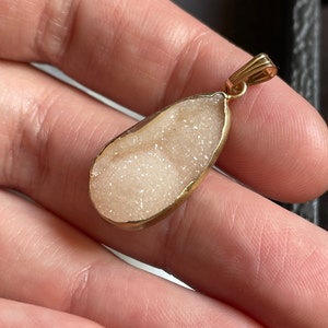 Puede incluir: Un colgante en forma de l&aacute;grima con un marco de metal dorado y una piedra blanca, brillante, similar a un cristal.