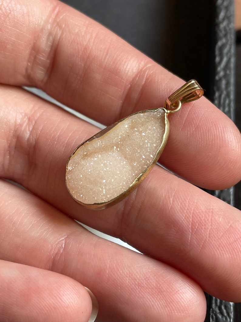 Puede incluir: Un colgante en forma de l&aacute;grima con un marco de metal dorado y una piedra blanca, brillante, similar a un cristal.