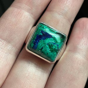K&ouml;nnte beinhalten: Ein quadratischer Ring mit einer polierten Silberschiene und einem eingefassten Edelstein, der eine marmorierte Oberfl&auml;che aus leuchtend gr&uuml;ner Malachit- und tiefblauer Azurit-Maserung aufweist. Der Stein zeigt nat&uuml;rliche Aderungen und Farbverl&auml;ufe.