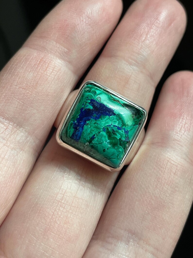 K&ouml;nnte beinhalten: Ein quadratischer Ring mit einer polierten Silberschiene und einem eingefassten Edelstein, der eine marmorierte Oberfl&auml;che aus leuchtend gr&uuml;ner Malachit- und tiefblauer Azurit-Maserung aufweist. Der Stein zeigt nat&uuml;rliche Aderungen und Farbverl&auml;ufe.