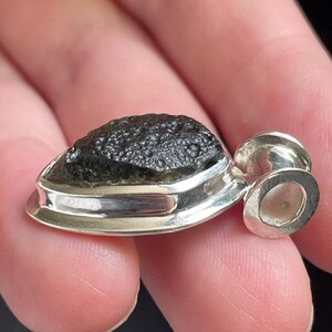May include: A pendant featuring a dark, textured, olive-green moldavite stone set in a polished silver bezel. The stone has a rough, pitted surface characteristic of natural tektite, while the silver setting includes a circular bail at the top.