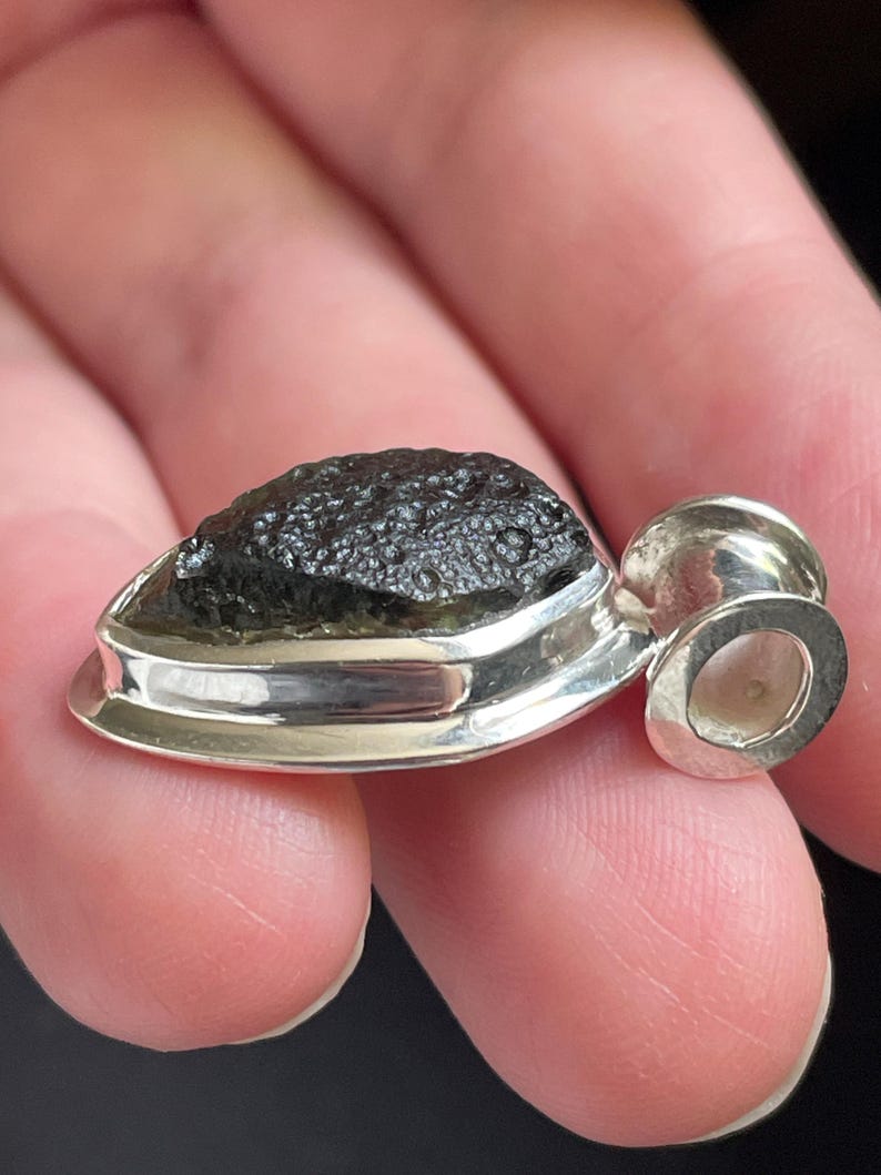 May include: A pendant featuring a dark, textured, olive-green moldavite stone set in a polished silver bezel. The stone has a rough, pitted surface characteristic of natural tektite, while the silver setting includes a circular bail at the top.